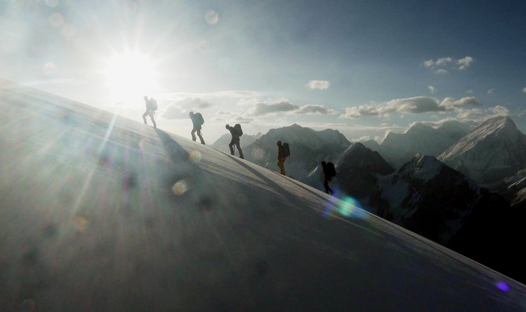 Fünf Bergsteiger gehen Berghang hinauf, die Sonne scheint in die Kamera