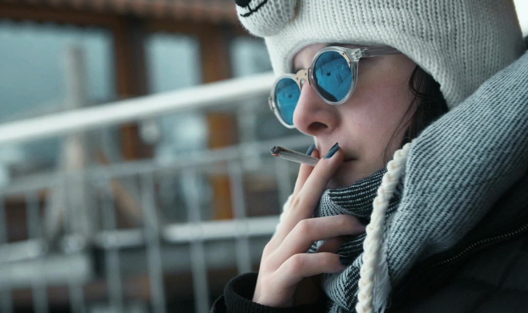 Magda-Lena sitzt auf der Terrasse und raucht. Sie trägt eine dicke Jacke Kappe und Sonnenbrille.