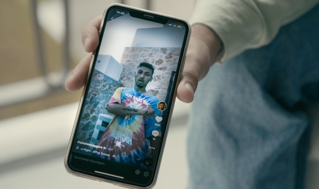 Ein Handy wird ins Bild gehalten mit einem laufenden Video von Younes Saggara.
