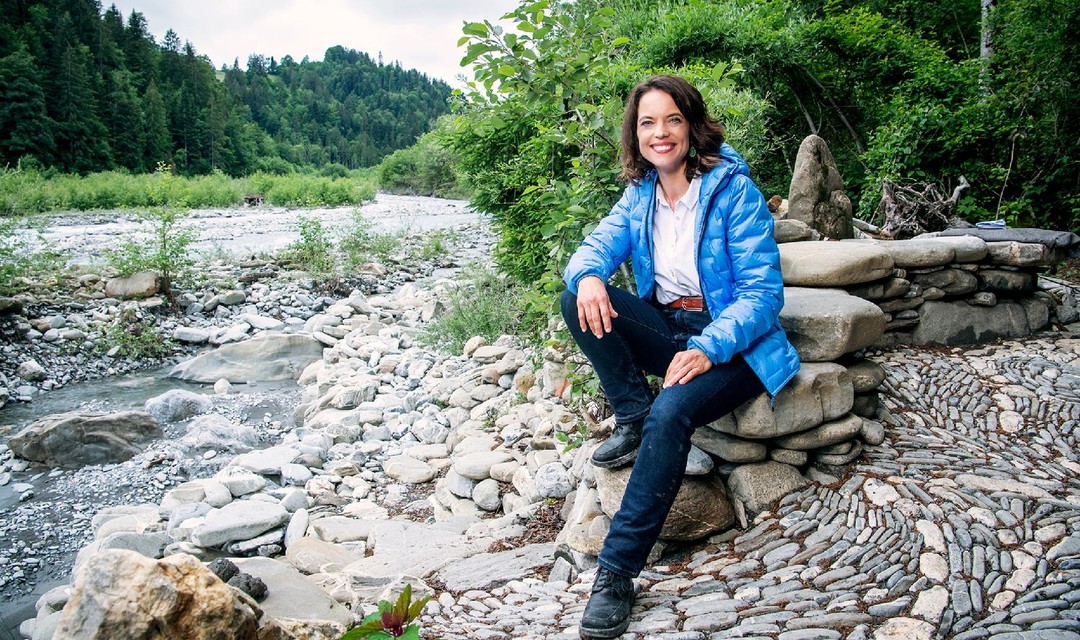 Mona Vetsch sitzt auf einem Steinhaufen an einem Fluss und lächelt in die Kamera