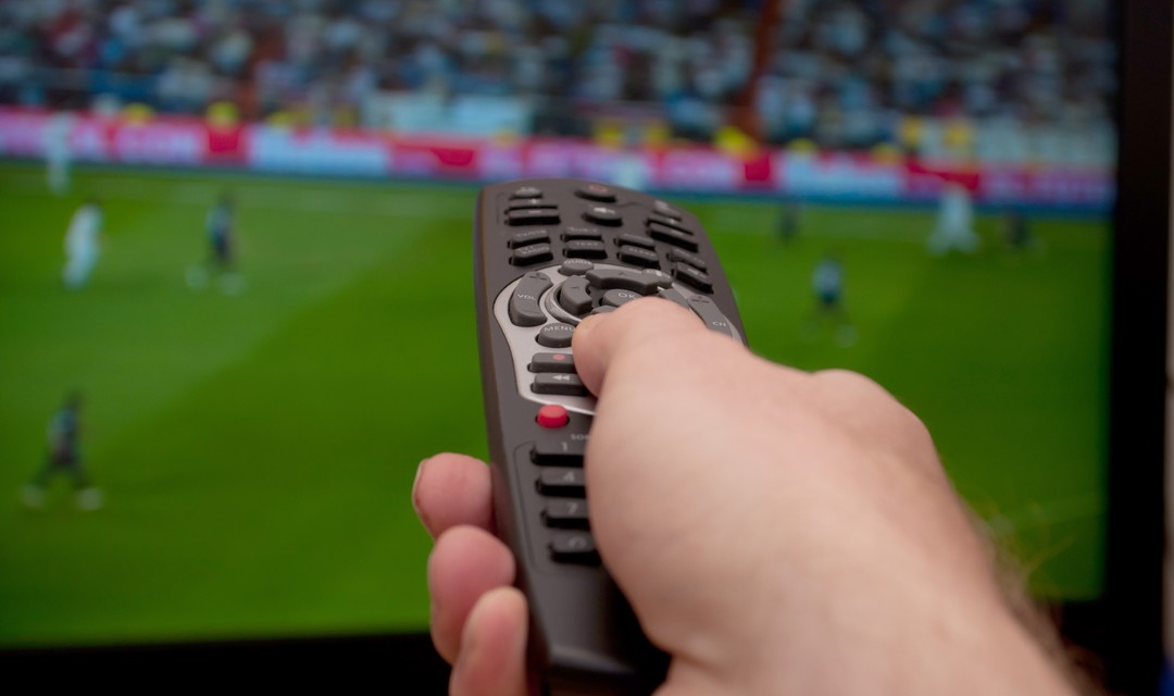 Fernbedienung zeigt auf Fernseher, auf welchem verschwommen ein Fussballspiel zu sehen ist.