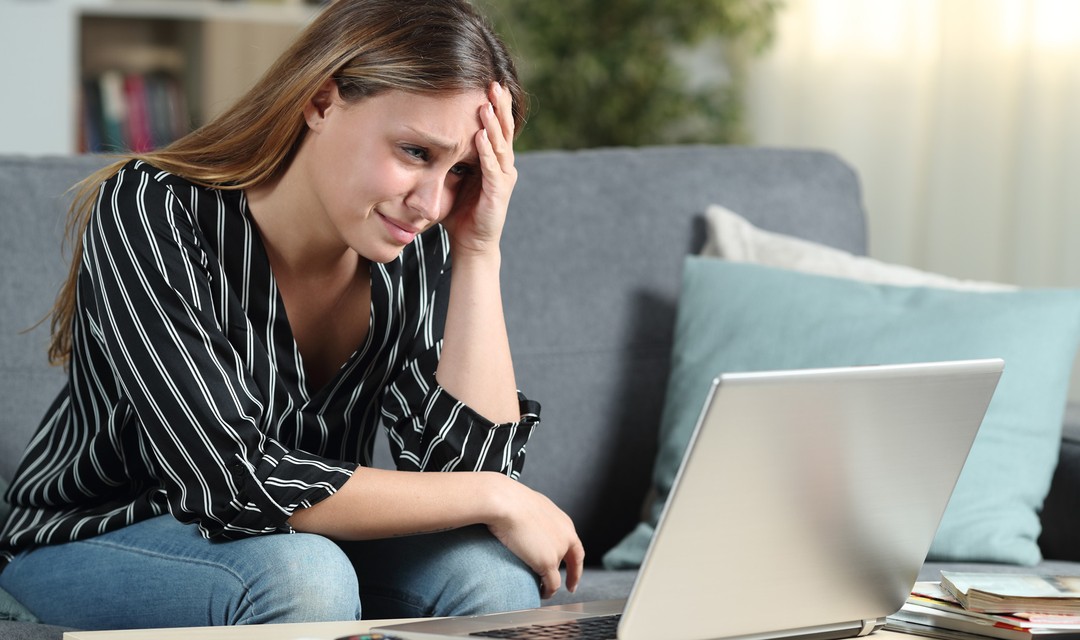 Eine junge Frau schaut zuhause ziemlich verzweifelt auf ihren Laptop-Bildschirm